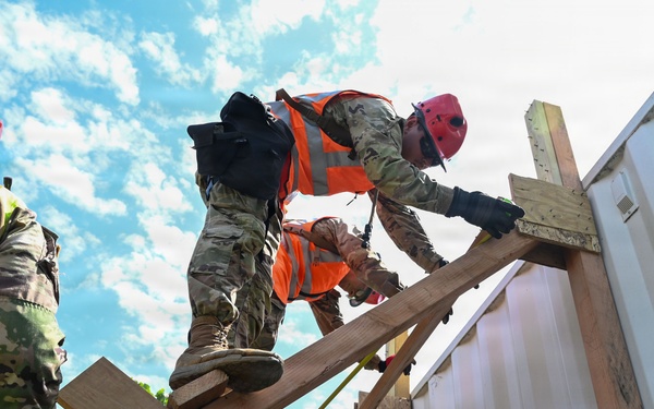 A group of military personnel in camouflage uniforms are working on a wooden structure, likely during a training exercise or disaster response operation. They are wearing high-visibility orange safety vests and red helmets while handling lumber on what appears to be a partially constructed or damaged wooden frame. The low-angle shot emphasizes the height and physical nature of the work being performed.