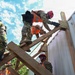 A group of military personnel in camouflage uniforms are working on a wooden structure, likely during a training exercise or disaster response operation. They are wearing high-visibility orange safety vests and red helmets while handling lumber on what appears to be a partially constructed or damaged wooden frame. The low-angle shot emphasizes the height and physical nature of the work being performed.