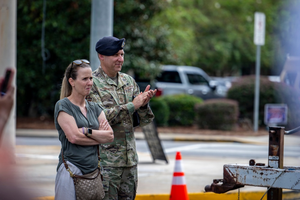 A military officer in US Army OCP (Operational Camouflage Pattern) uniform and dark blue beret stands alongside a civilian woman in a gray top. The officer appears to be clapping or applauding while the woman stands with her arms crossed. They are outdoors in what appears to be a military installation or base environment.