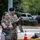 A military officer in US Army OCP (Operational Camouflage Pattern) uniform and dark blue beret stands alongside a civilian woman in a gray top. The officer appears to be clapping or applauding while the woman stands with her arms crossed. They are outdoors in what appears to be a military installation or base environment.
