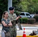 A military officer in US Army OCP (Operational Camouflage Pattern) uniform and dark blue beret stands alongside a civilian woman in a gray top. The officer appears to be clapping or applauding while the woman stands with her arms crossed. They are outdoors in what appears to be a military installation or base environment.