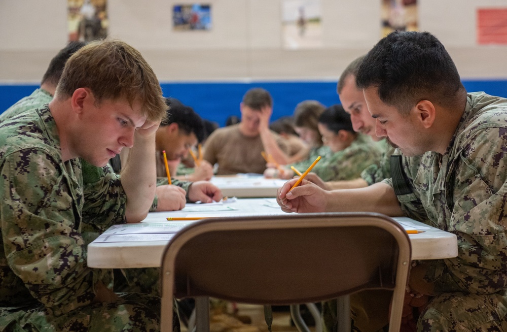 A group of military personnel in camouflage uniforms are seated at tables in what appears to be a gymnasium or training facility, engaged in a written examination or assessment. The foreground shows two service members leaning over papers with pencils in hand, with additional personnel visible in the background doing the same. The uniforms appear consistent with U.S. Navy digital camouflage (NWU Type I pattern).