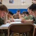 A group of military personnel in camouflage uniforms are seated at tables in what appears to be a gymnasium or training facility, engaged in a written examination or assessment. The foreground shows two service members leaning over papers with pencils in hand, with additional personnel visible in the background doing the same. The uniforms appear consistent with U.S. Navy digital camouflage (NWU Type I pattern).