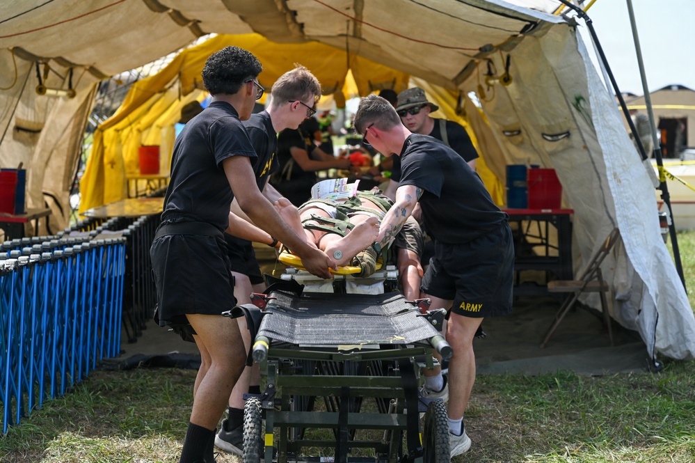 Several U.S. Army personnel dressed in black PT (physical training) uniforms are transporting a patient on a wheeled stretcher/gurney into a large military field tent. The scene appears to be a medical training exercise or field hospital simulation, with additional personnel visible working inside the tent. A simulated or real casualty is strapped to the stretcher with visible medical equipment attached.