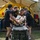Several U.S. Army personnel dressed in black PT (physical training) uniforms are transporting a patient on a wheeled stretcher/gurney into a large military field tent. The scene appears to be a medical training exercise or field hospital simulation, with additional personnel visible working inside the tent. A simulated or real casualty is strapped to the stretcher with visible medical equipment attached.