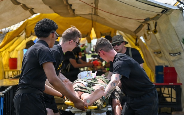 Several U.S. Army personnel dressed in black PT (physical training) uniforms are transporting a patient on a wheeled stretcher/gurney into a large military field tent. The scene appears to be a medical training exercise or field hospital simulation, with additional personnel visible working inside the tent. A simulated or real casualty is strapped to the stretcher with visible medical equipment attached.