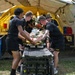 Several U.S. Army personnel dressed in black PT (physical training) uniforms are transporting a patient on a wheeled stretcher/gurney into a large military field tent. The scene appears to be a medical training exercise or field hospital simulation, with additional personnel visible working inside the tent. A simulated or real casualty is strapped to the stretcher with visible medical equipment attached.