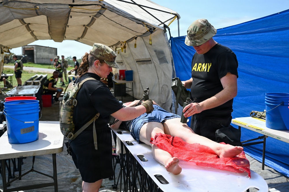 Two U.S. Army personnel in military uniforms are conducting a medical training exercise under a field tent. They are treating a simulated casualty mannequin on a stretcher table, with one applying a tourniquet to the leg area. The setting appears to be an outdoor military training environment with additional personnel visible in the background.