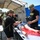 Two U.S. Army personnel in military uniforms are conducting a medical training exercise under a field tent. They are treating a simulated casualty mannequin on a stretcher table, with one applying a tourniquet to the leg area. The setting appears to be an outdoor military training environment with additional personnel visible in the background.