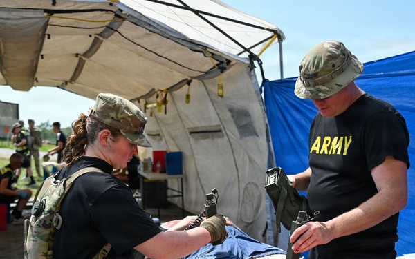 Two U.S. Army personnel in military uniforms are conducting a medical training exercise under a field tent. They are treating a simulated casualty mannequin on a stretcher table, with one applying a tourniquet to the leg area. The setting appears to be an outdoor military training environment with additional personnel visible in the background.