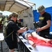 Two U.S. Army personnel in military uniforms are conducting a medical training exercise under a field tent. They are treating a simulated casualty mannequin on a stretcher table, with one applying a tourniquet to the leg area. The setting appears to be an outdoor military training environment with additional personnel visible in the background.