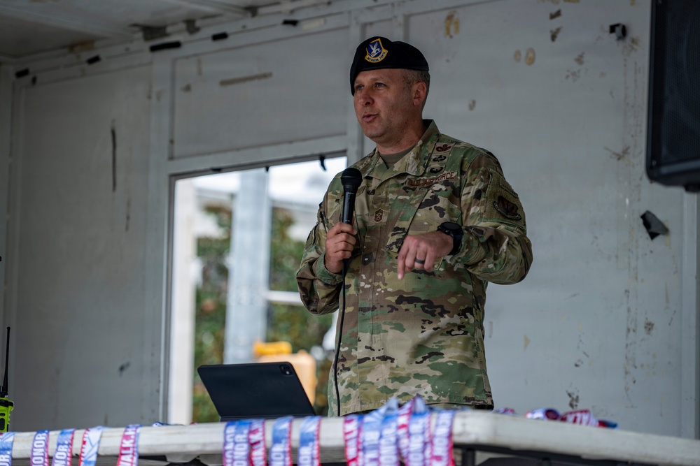 A U.S. Air Force military personnel member is speaking at a podium or table while holding a microphone. He is wearing OCP (Operational Camouflage Pattern) uniform with a black beret bearing a military insignia. The setting appears to be an outdoor or semi-covered venue, with what appear to be event lanyards or medals on the table in front of him.