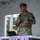 A U.S. Air Force military personnel member is speaking at a podium or table while holding a microphone. He is wearing OCP (Operational Camouflage Pattern) uniform with a black beret bearing a military insignia. The setting appears to be an outdoor or semi-covered venue, with what appear to be event lanyards or medals on the table in front of him.