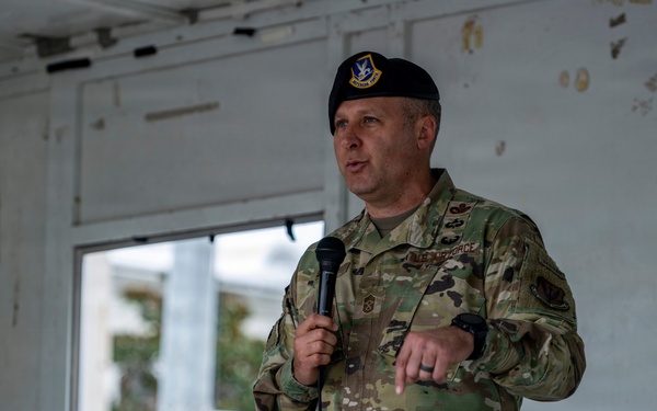 A U.S. Air Force military personnel member is speaking at a podium or table while holding a microphone. He is wearing OCP (Operational Camouflage Pattern) uniform with a black beret bearing a military insignia. The setting appears to be an outdoor or semi-covered venue, with what appear to be event lanyards or medals on the table in front of him.