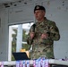 A U.S. Air Force military personnel member is speaking at a podium or table while holding a microphone. He is wearing OCP (Operational Camouflage Pattern) uniform with a black beret bearing a military insignia. The setting appears to be an outdoor or semi-covered venue, with what appear to be event lanyards or medals on the table in front of him.