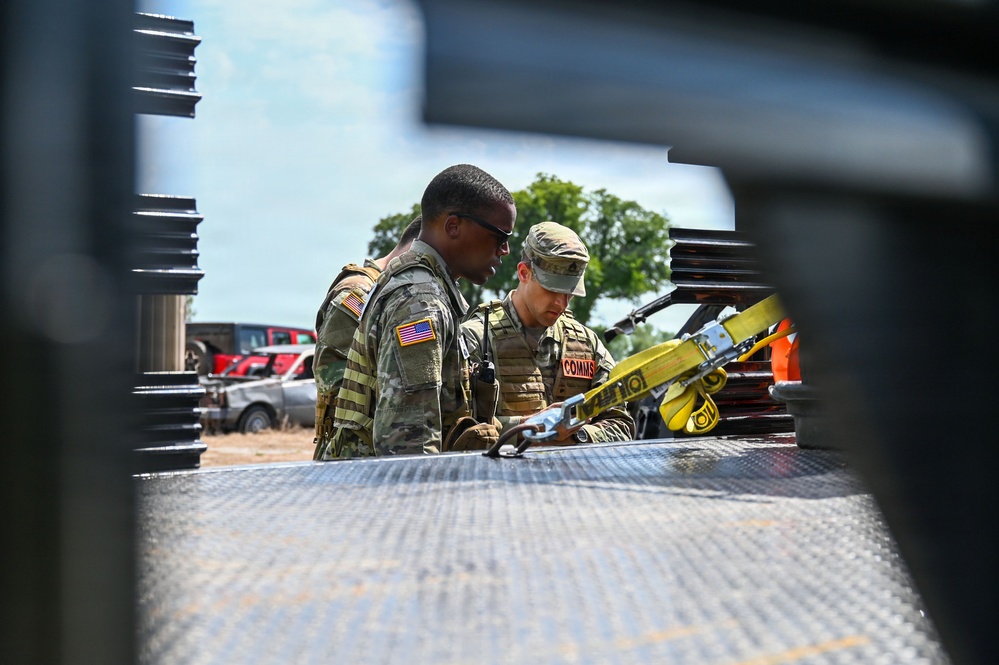 Two U.S. Army soldiers in OCP (Operational Camouflage Pattern) uniforms are working together near the bed of a military or utility vehicle. One soldier wears sunglasses and displays a U.S. flag patch on their left shoulder, while the other has a 'COMMS' identifier patch visible on their vest. They appear to be inspecting or securing equipment, including yellow ratchet straps, on a diamond-plate metal surface.
