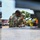 Two U.S. Army soldiers in OCP (Operational Camouflage Pattern) uniforms are working together near the bed of a military or utility vehicle. One soldier wears sunglasses and displays a U.S. flag patch on their left shoulder, while the other has a 'COMMS' identifier patch visible on their vest. They appear to be inspecting or securing equipment, including yellow ratchet straps, on a diamond-plate metal surface.