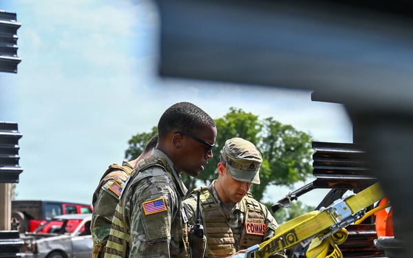 Two U.S. Army soldiers in OCP (Operational Camouflage Pattern) uniforms are working together near the bed of a military or utility vehicle. One soldier wears sunglasses and displays a U.S. flag patch on their left shoulder, while the other has a 'COMMS' identifier patch visible on their vest. They appear to be inspecting or securing equipment, including yellow ratchet straps, on a diamond-plate metal surface.