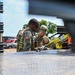 Two U.S. Army soldiers in OCP (Operational Camouflage Pattern) uniforms are working together near the bed of a military or utility vehicle. One soldier wears sunglasses and displays a U.S. flag patch on their left shoulder, while the other has a 'COMMS' identifier patch visible on their vest. They appear to be inspecting or securing equipment, including yellow ratchet straps, on a diamond-plate metal surface.
