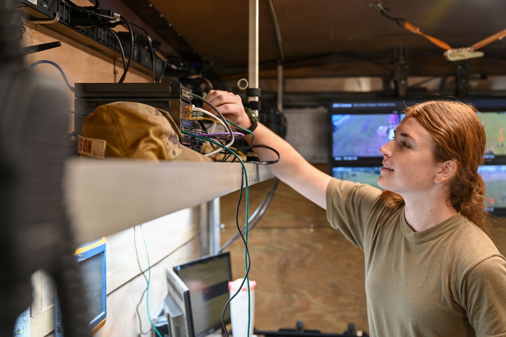 A person with long red hair wearing an olive/tan military-style t-shirt is working on electronic equipment mounted on a shelf inside what appears to be a mobile command or communications vehicle. They are adjusting or connecting cables and components on a rack-mounted system. Multiple monitors and screens are visible in the background, suggesting a tactical or operational environment.
