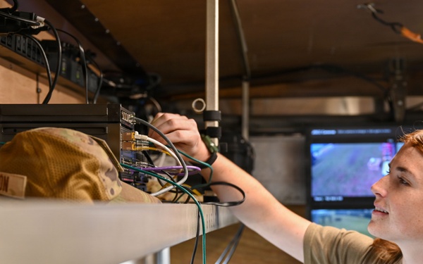 A person with long red hair wearing an olive/tan military-style t-shirt is working on electronic equipment mounted on a shelf inside what appears to be a mobile command or communications vehicle. They are adjusting or connecting cables and components on a rack-mounted system. Multiple monitors and screens are visible in the background, suggesting a tactical or operational environment.