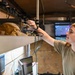 A person with long red hair wearing an olive/tan military-style t-shirt is working on electronic equipment mounted on a shelf inside what appears to be a mobile command or communications vehicle. They are adjusting or connecting cables and components on a rack-mounted system. Multiple monitors and screens are visible in the background, suggesting a tactical or operational environment.