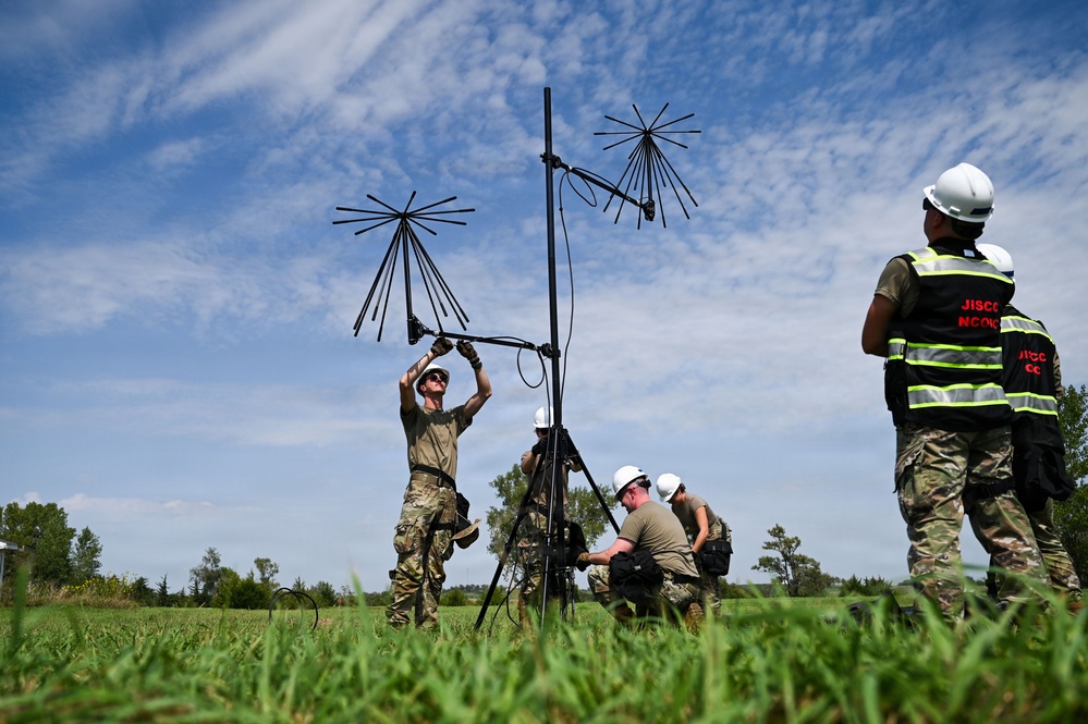 Military personnel in camouflage uniforms are assembling or setting up antenna equipment on a tripod in an open grassy field. Two individuals wearing safety vests labeled 'JISCC NCOIC' and white hard hats are observing the setup process. The scene appears to be a field training or communications equipment deployment exercise.