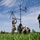 Military personnel in camouflage uniforms are assembling or setting up antenna equipment on a tripod in an open grassy field. Two individuals wearing safety vests labeled 'JISCC NCOIC' and white hard hats are observing the setup process. The scene appears to be a field training or communications equipment deployment exercise.