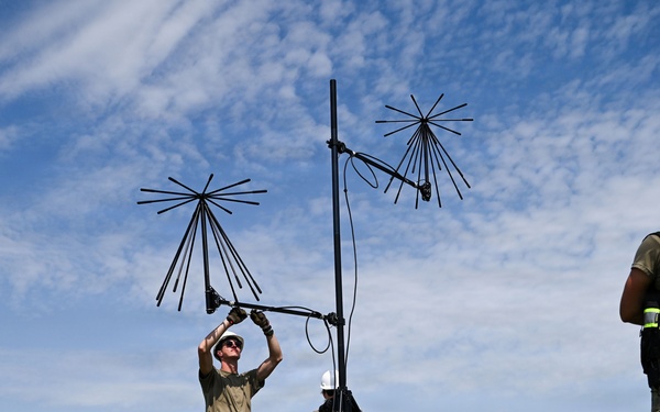 Military personnel in camouflage uniforms are assembling or setting up antenna equipment on a tripod in an open grassy field. Two individuals wearing safety vests labeled 'JISCC NCOIC' and white hard hats are observing the setup process. The scene appears to be a field training or communications equipment deployment exercise.