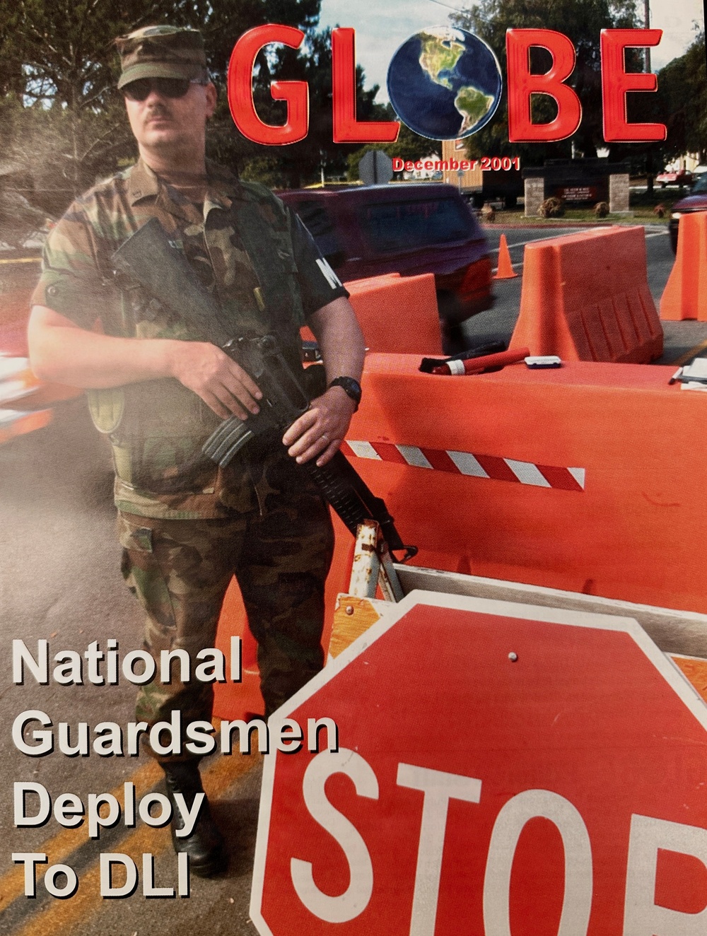 This is the December 2001 cover of 'GLOBE' magazine, featuring a National Guardsman in woodland camouflage BDUs standing at a security checkpoint. The cover story headline reads 'National Guardsmen Deploy To DLI' (Defense Language Institute), likely reflecting post-9/11 heightened security measures at military installations. The soldier is armed and positioned near orange traffic barriers and a stop sign at what appears to be a base entry control point.