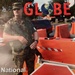 This is the December 2001 cover of 'GLOBE' magazine, featuring a National Guardsman in woodland camouflage BDUs standing at a security checkpoint. The cover story headline reads 'National Guardsmen Deploy To DLI' (Defense Language Institute), likely reflecting post-9/11 heightened security measures at military installations. The soldier is armed and positioned near orange traffic barriers and a stop sign at what appears to be a base entry control point.