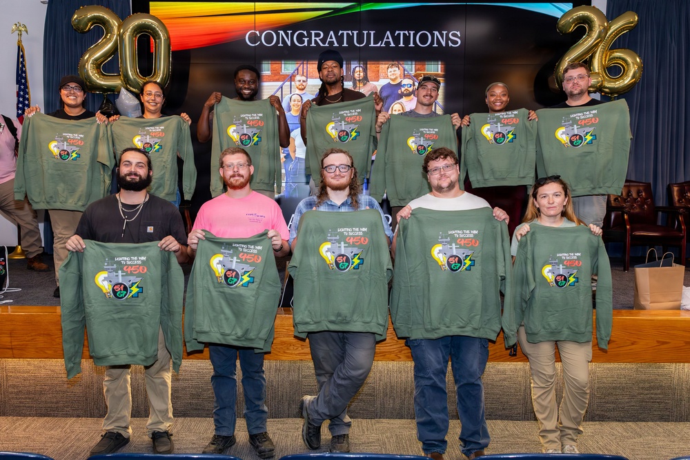 A group of approximately 12 people pose for a congratulatory photo, each holding up olive/army green sweatshirts with a graphic design reading 'Lighting the Way to Success 450' with what appears to be an electrician/utility themed logo. Gold number balloons spelling '2026' flank a screen displaying 'CONGRATULATIONS' in the background. The event appears to be a graduation or program completion ceremony.