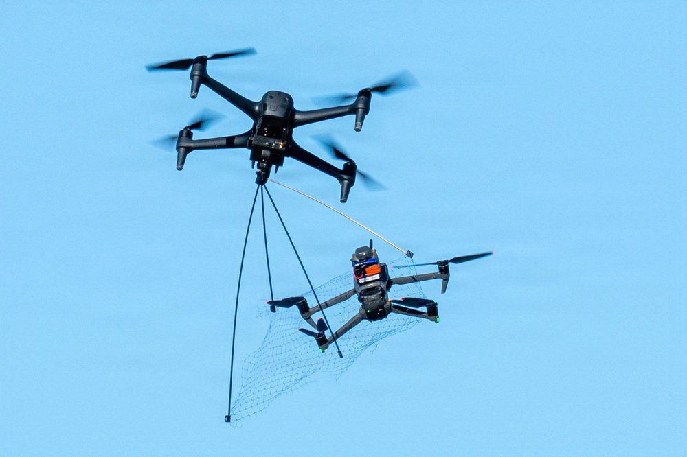 A large black quadcopter drone is airborne and has deployed a net to capture a smaller quadcopter drone below it. The smaller drone is entangled in the net, which is suspended by multiple tethering lines from the larger interceptor drone. This appears to be a counter-drone or drone interdiction system in operation.