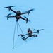 A large black quadcopter drone is airborne and has deployed a net to capture a smaller quadcopter drone below it. The smaller drone is entangled in the net, which is suspended by multiple tethering lines from the larger interceptor drone. This appears to be a counter-drone or drone interdiction system in operation.
