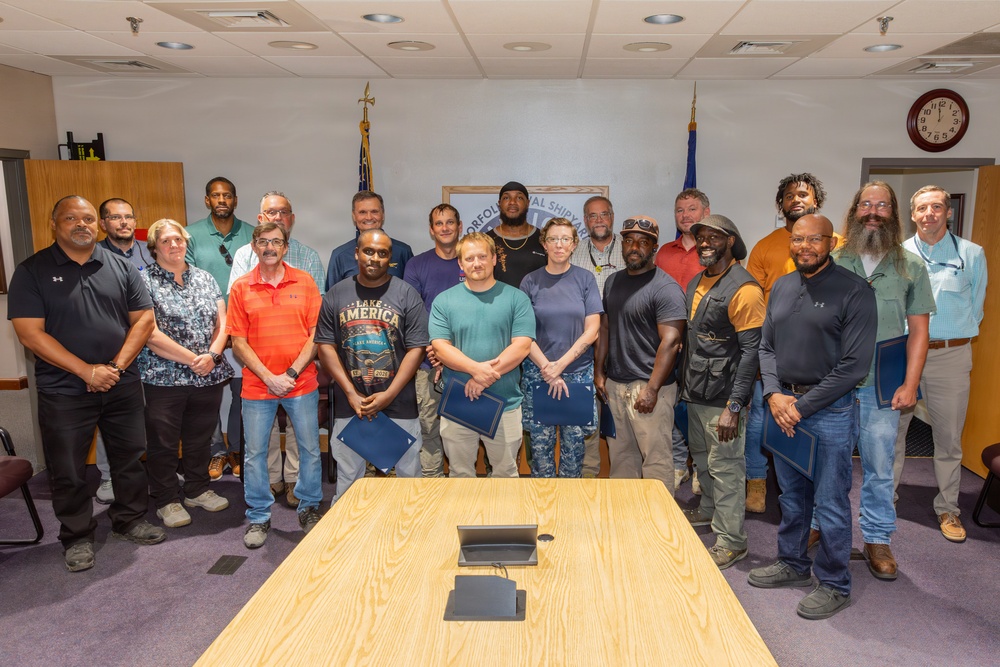 A group of approximately 20 people pose for a formal group photo in what appears to be a conference room at Norfolk Naval Shipyard, as indicated by the partially visible sign in the background. Several individuals in the front row are holding blue folders or certificates, suggesting this is a recognition or award ceremony. The setting includes American and Navy flags flanking the organizational sign.