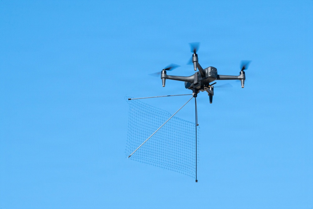 A dark gray quadcopter drone is captured mid-flight against a clear blue sky. The drone is carrying a suspended net apparatus attached via rigid rods, suggesting it is configured for intercepting or capturing another drone or object. The image is sharp and well-lit, providing excellent detail of both the drone and its payload.