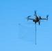 A dark gray quadcopter drone is captured mid-flight against a clear blue sky. The drone is carrying a suspended net apparatus attached via rigid rods, suggesting it is configured for intercepting or capturing another drone or object. The image is sharp and well-lit, providing excellent detail of both the drone and its payload.