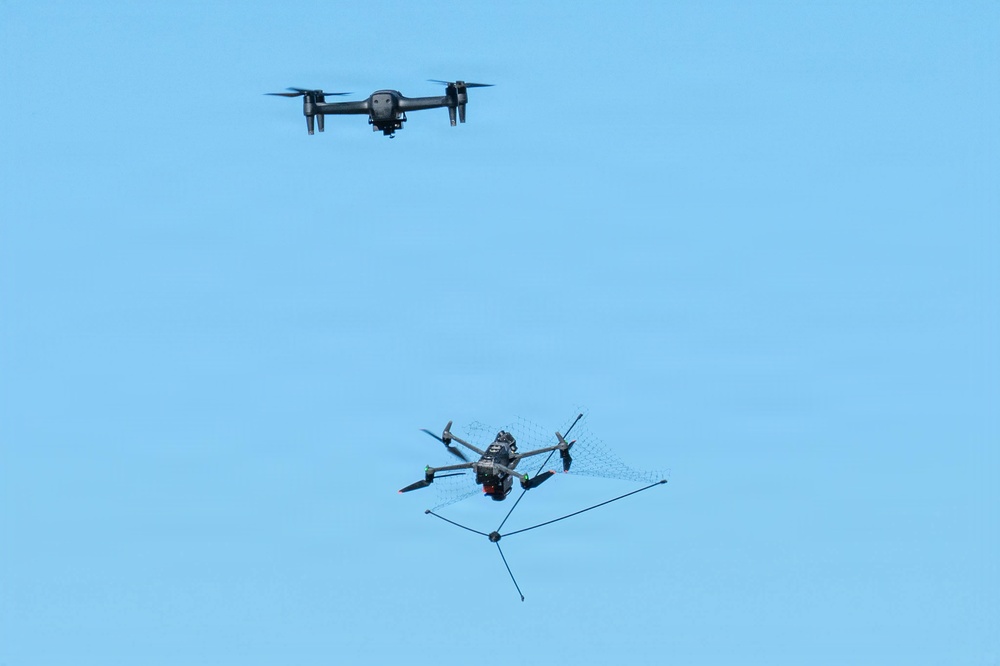 The image shows two drones in flight against a clear blue sky. The lower drone appears to be a counter-drone or interceptor unit equipped with a net-launching or net-catching mechanism, while the upper drone appears to be a standard quadcopter, possibly the target. This appears to depict a drone interception or counter-UAS (unmanned aerial system) operation.