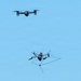 The image shows two drones in flight against a clear blue sky. The lower drone appears to be a counter-drone or interceptor unit equipped with a net-launching or net-catching mechanism, while the upper drone appears to be a standard quadcopter, possibly the target. This appears to depict a drone interception or counter-UAS (unmanned aerial system) operation.