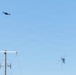 The image shows two unmanned aerial vehicles (drones) flying in a clear blue sky above a utility power pole with electrical insulators and wires. One drone in the upper left appears to be a standard quadcopter, while the second drone on the right has a more complex, multi-legged or interceptor-style frame and appears to be in a tilted or dynamic flight posture. The scene suggests a possible drone interception or counter-drone operation scenario.