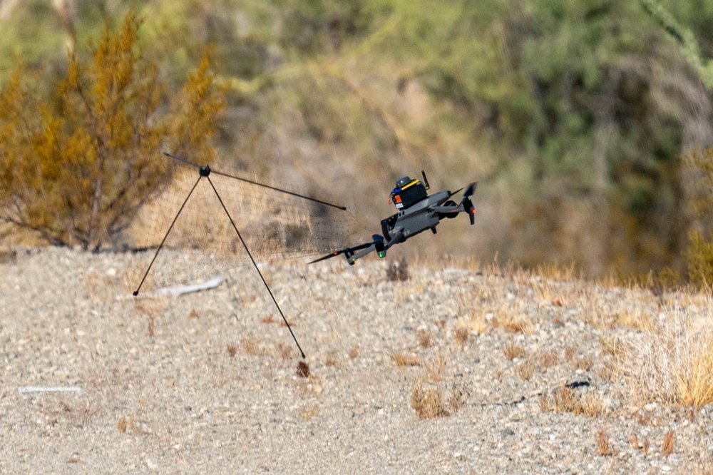 A consumer-grade quadcopter drone (appears to be a DJI Mavic series) is captured mid-flight in an arid, desert-scrub environment. The drone has an additional payload or sensor module mounted on top and is entangled with or deploying a net attached to a collapsible frame structure. The scene suggests either a counter-drone net-capture system test or a drone-deployed net system in operation.