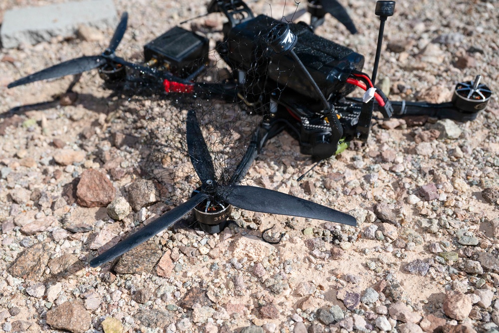 A crashed or downed FPV racing drone lies on a rocky, gravelly surface. The drone appears to have been entangled in a net or mesh material, which is wrapped around its frame and propellers. The drone is black with red wiring visible, and one detached motor with a tri-blade propeller is separated from the main frame in the foreground.