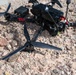 A crashed or downed FPV racing drone lies on a rocky, gravelly surface. The drone appears to have been entangled in a net or mesh material, which is wrapped around its frame and propellers. The drone is black with red wiring visible, and one detached motor with a tri-blade propeller is separated from the main frame in the foreground.