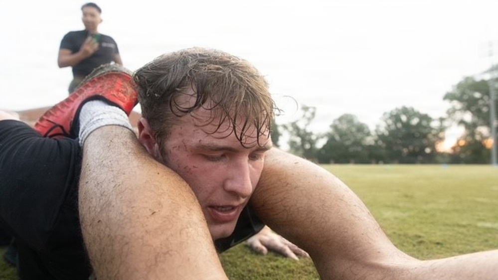 A close-up action shot of a rugby player on the ground, appearing to be in a tackle or ruck situation during a match or training session. The player is visibly muddy and wet, suggesting play in damp conditions. A second person in a black shirt is visible standing in the background on a grass field.