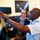 Two men are examining multiple computer monitors in what appears to be a military or aerospace operations center. A uniformed military officer is pointing at one of the screens while a civilian in a blazer observes. A satellite imagery poster is visible on the wall in the background.