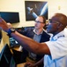 Two men are examining multiple computer monitors in what appears to be a military or aerospace operations center. A uniformed military officer is pointing at one of the screens while a civilian in a blazer observes. A satellite imagery poster is visible on the wall in the background.
