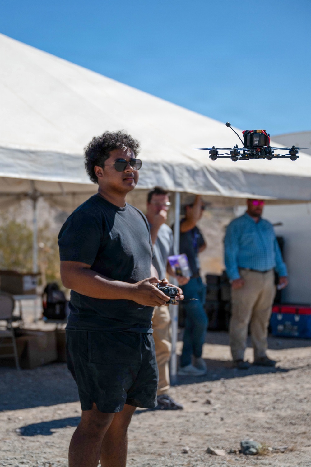 A man wearing sunglasses and a dark t-shirt is operating a quadcopter drone using a handheld remote controller in an outdoor desert-like setting. A racing-style FPV drone with a camera mount is visible hovering in the upper right of the frame. Several other people are visible in the background under a large white canopy tent.