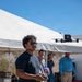 A man wearing sunglasses and a dark t-shirt is operating a quadcopter drone using a handheld remote controller in an outdoor desert-like setting. A racing-style FPV drone with a camera mount is visible hovering in the upper right of the frame. Several other people are visible in the background under a large white canopy tent.