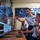 Three uniformed U.S. Air Force personnel are working at a computer workstation in what appears to be a military operations or cyber command center. One standing officer is pointing at a large monitor displaying data dashboards and code, while two seated airmen observe and operate the equipment. The background features a large display wall with multiple screens showing space-related imagery and data.