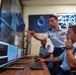 Three uniformed U.S. Air Force personnel are working at a computer workstation in what appears to be a military operations or cyber command center. One standing officer is pointing at a large monitor displaying data dashboards and code, while two seated airmen observe and operate the equipment. The background features a large display wall with multiple screens showing space-related imagery and data.